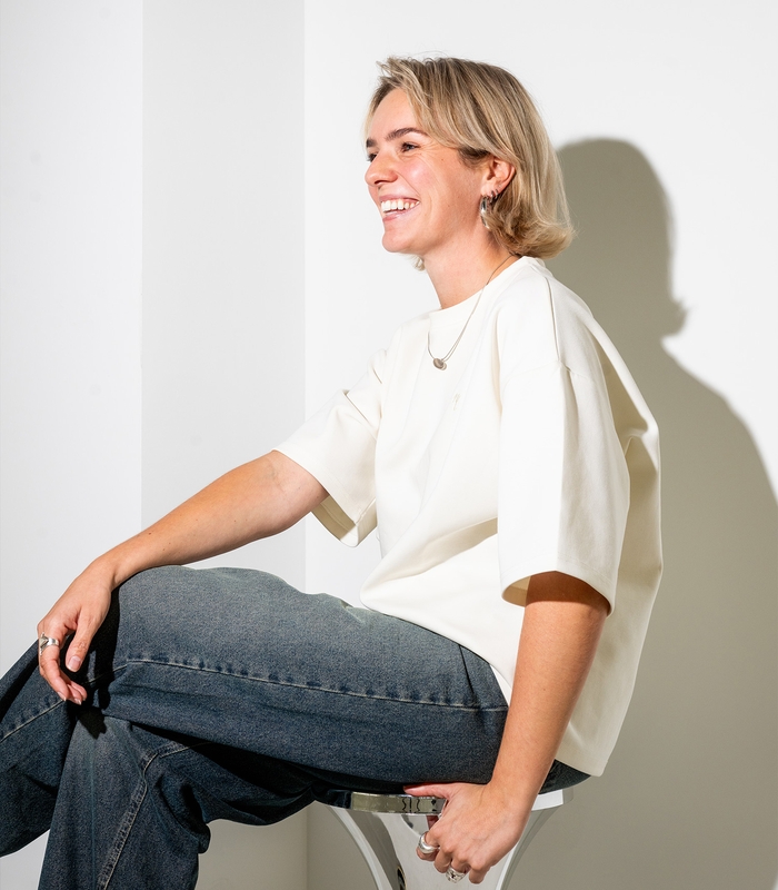 Persoon met kort blond haar lacht, zittend op een stoel. Ze draagt een wit T-shirt en een spijkerbroek. Schaduw op witte muur.
