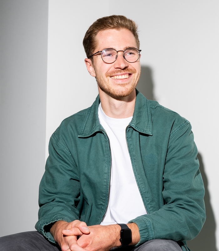 Man met bril glimlacht, draagt een groene jas en witte T-shirt, handen gevouwen op schoot, zittend tegen een witte muur.