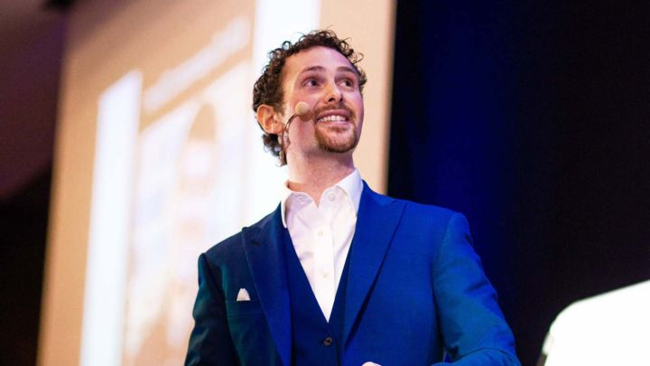 Smiling man in a blue suit and white shirt wearing a headset microphone, speaking on stage with a projected screen behind him.