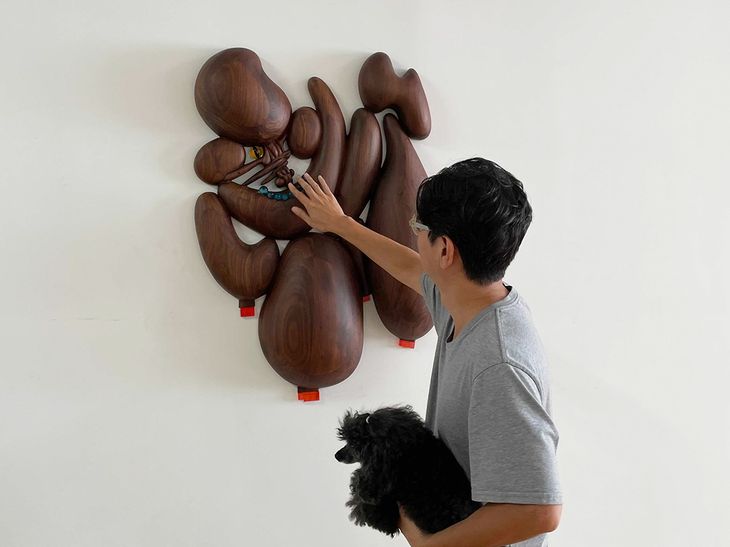 artist holding a black poodle, inspecting a wooden wall-mounted sculpture