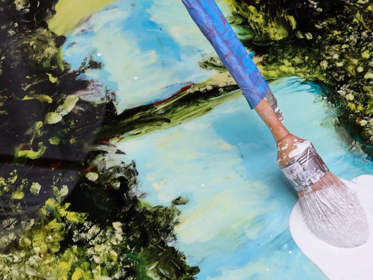 big white brush on a long wooden stick adding white oil paint to the surface of a large, glossy print