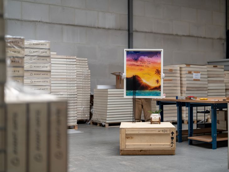 A figure holding up a white framed Friedrich Kunath print in a warehouse surrounded by boxes