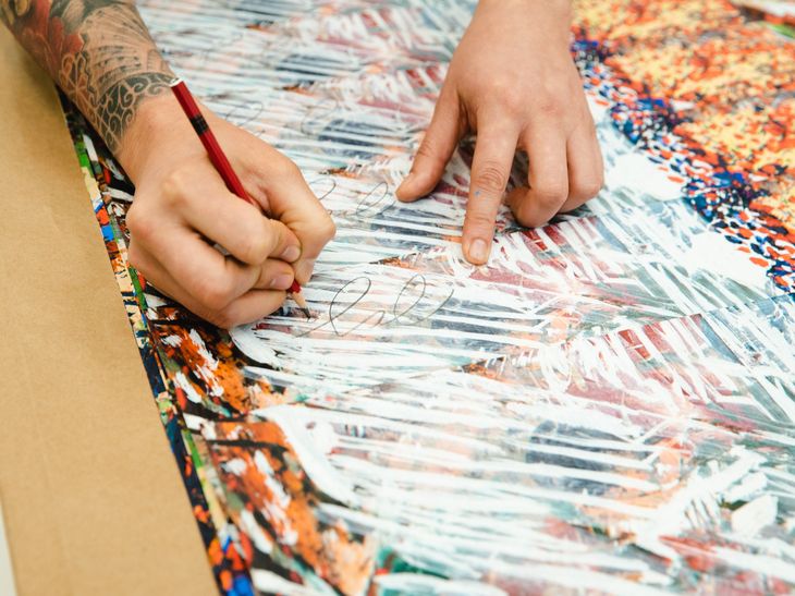 a pile of prints being signed by an artists, only his hands are visible and he is using a red pencil
