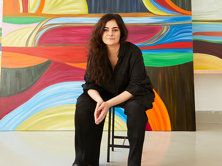 Marina Perez Simao sitting on a chair in front of several colourful paintings