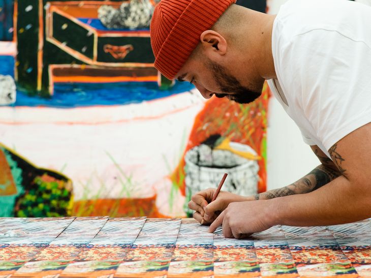 artist signing a stack of prints while wearing an orange beanie hat, a dark-toned painting is hanging on the wall behind him