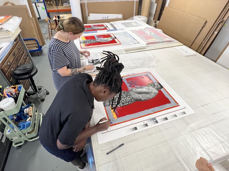 artist signing the bottom right corner of a large red artwork in a busy print workshop