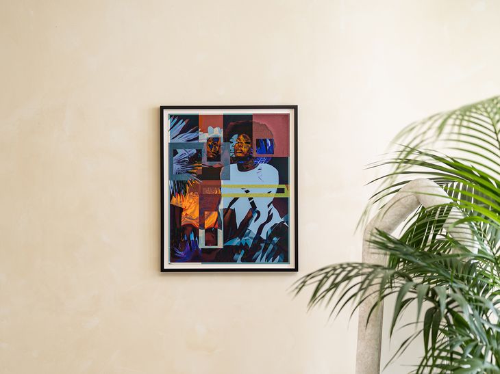 A framed print edition by Amani Lewis hung on a white wall above a chair and plant