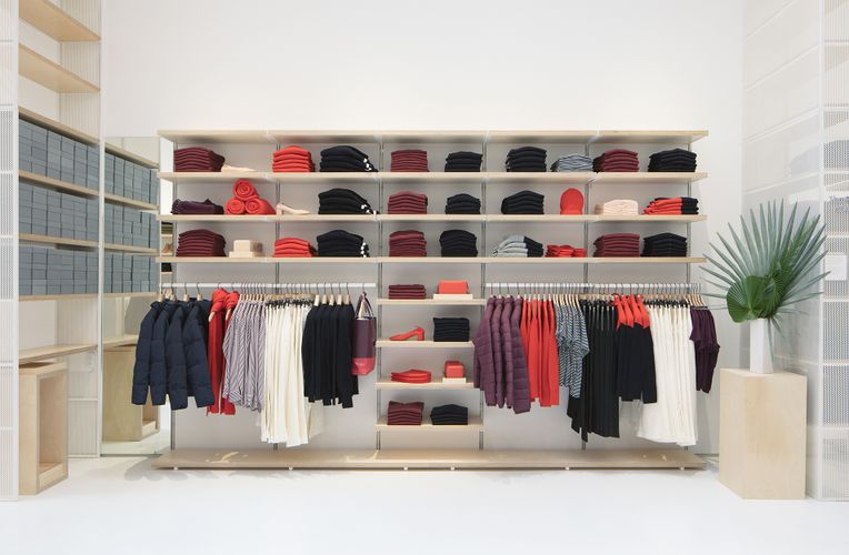 Wooden System 1224 shelves against a white wall are used to display various clothing items. More shelves are lined along the left wall holding boxes.