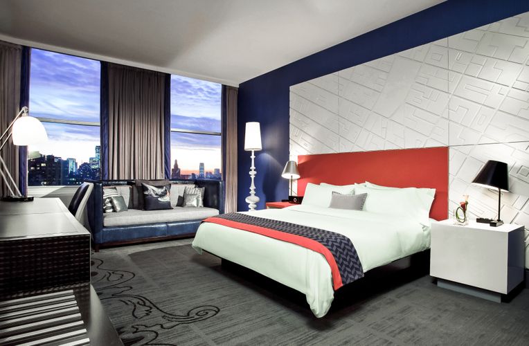 View of a bedroom space. The wall is navy ad white with geometric white Iconic Panel. A red bed frame is placed against the wall and the room is furnished with lights and a navy couch.