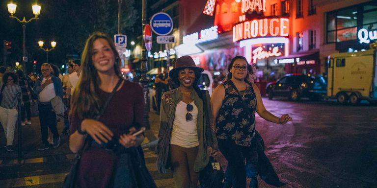 Three girls smiling, walking along a lit up European street at nighttime.
