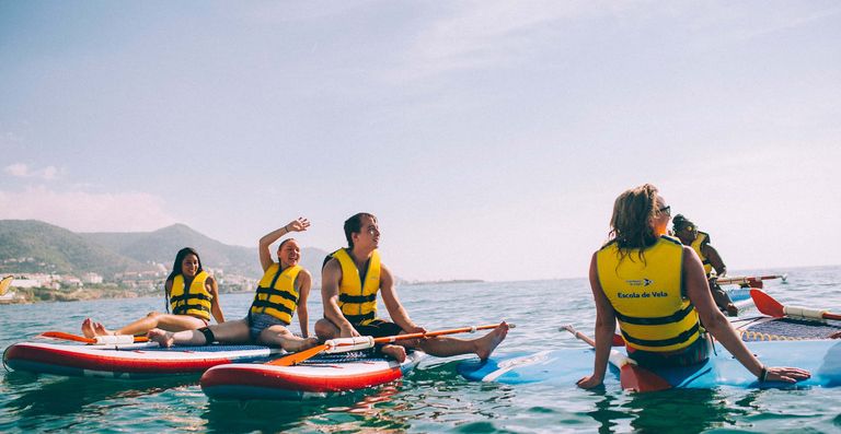 A group of travelers sitting on paddle boards in the ocean, smiling and laughing, somewhere in Europe with scenic houses in the background on shore.