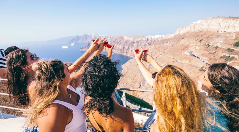 A group of women holding up wine glasses as they overlook a cliffside in Greece.