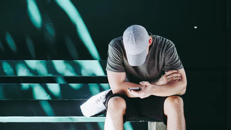 A man wearing a baseball cap sits on a green bench. He looks down at his phone.