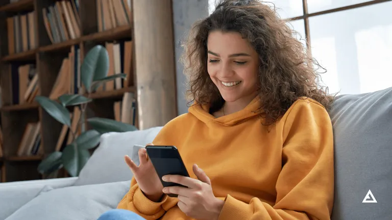 A woman wearing an orange hoodie sits on a grey couch and scrolls on her phone.