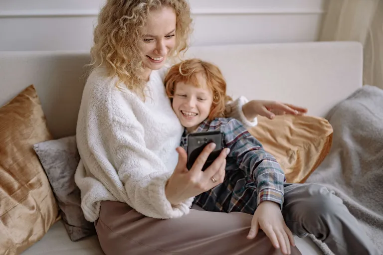 A mother and son sit on the couch smiling at a cellphone.