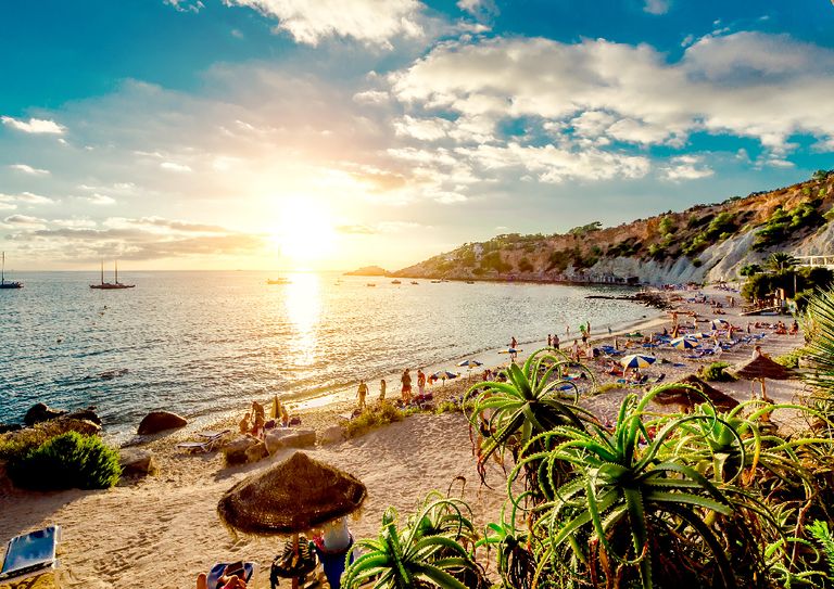A beach in Ibiza with the sunset over the ocean and people lounging on the sand.