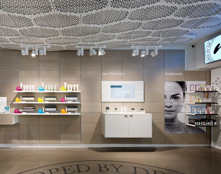 Interior of a beauty store. Various beauty products are lined up along white shelves against a beige wall.