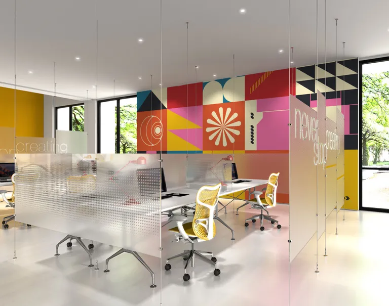 A colorfully decorated office space is divided up with frosted glass panels held up by thin metallic cables and rods. Within the spaces are desks with monitors and yellow rolling chairs.