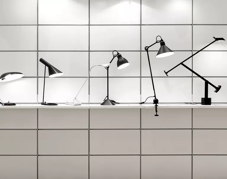 Various black lamps are lined up along a white shelf held up against a grid patterned wall.