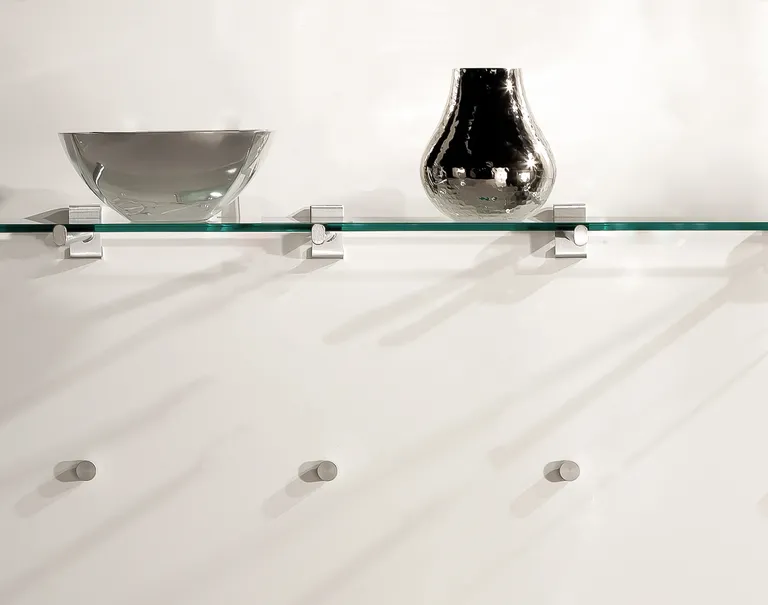 A glass shelf displaying a metal bowl and vase is propped up using the Puck System against a white wall.