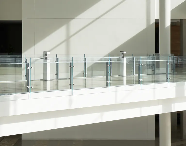 Wide shot of a hallway inside a large white building. Glass walls reinforce the hallway with two white hand sanitizing stations placed along the path.
