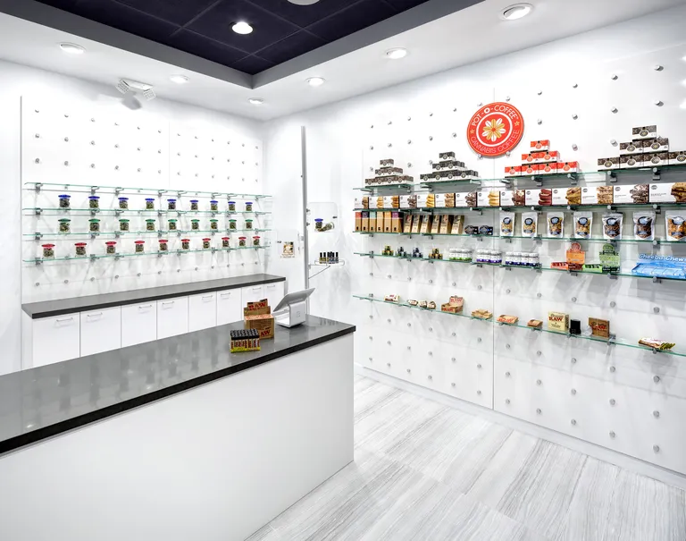 View of the interior of a white coffee shop. A white counter with a black top is placed in front of two white walls both utilizing the puck system. Glass shelves are held up against each wall displaying a variety of different snacks. Over the shelves, an orange circle with the logo for "Pot-O-Coffee. Cannabis Coffee" is hung.