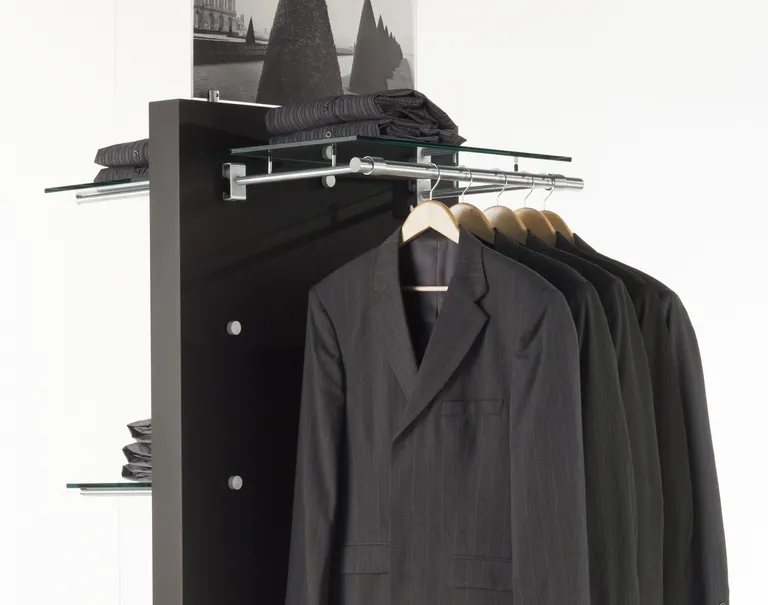 Various black clothing articles are on display against a black standing panel with metal Puck fixtures attached to it. The fixtures hold up metal bars and glass shelves for the clothes to be displayed on.