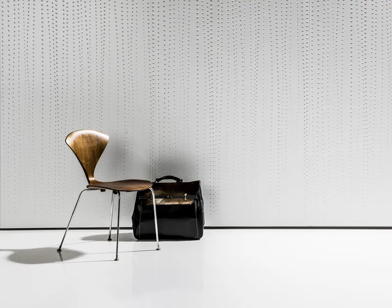 A wooden chair with a black suitcase are propped in front of a white wall decorated in dotted Iconic Panel.