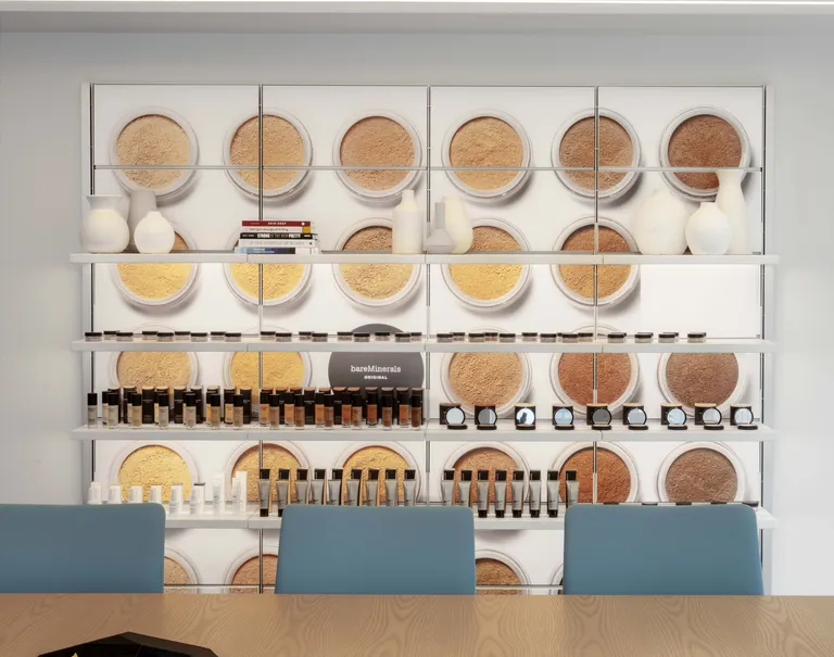 View of a wall decorated with lit up panels with an image of different foundation powders. The panels are equipped with white System 1224 shelves showcasing miscellaneous items including makeup products, books, and vases.