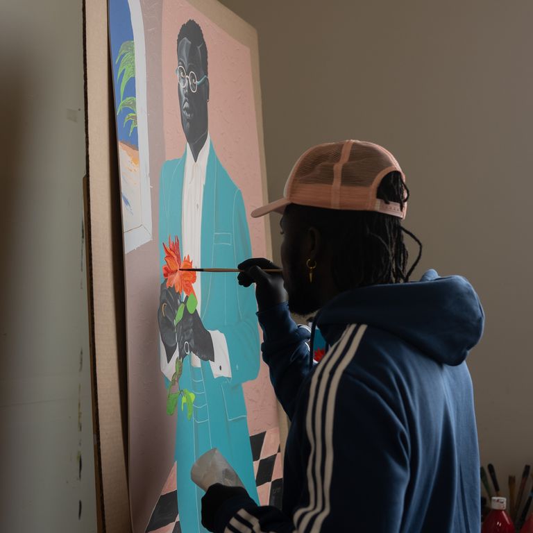 artist Otis Kwame Kye Quaicoe hand holding a paintbrush up to a painting to add detail to an orange flower
