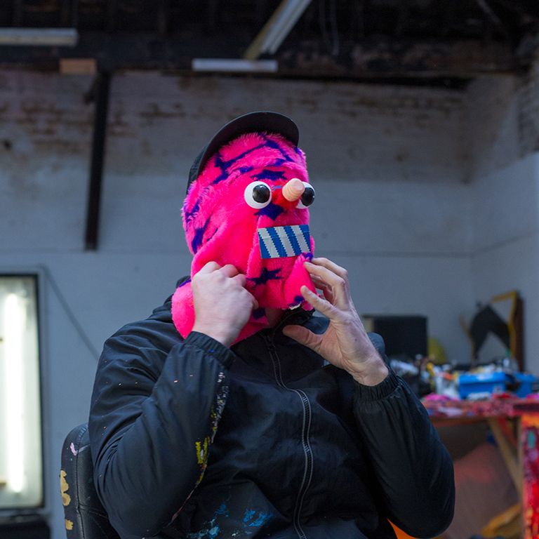 artist wearing black jacket with his hands clutching a pink mask which covers his face