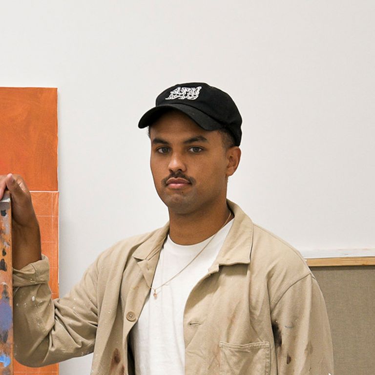 headshot of artist wearing white t-shirt, beige jacket and a black cap as he rests his hands on an easel in his studio