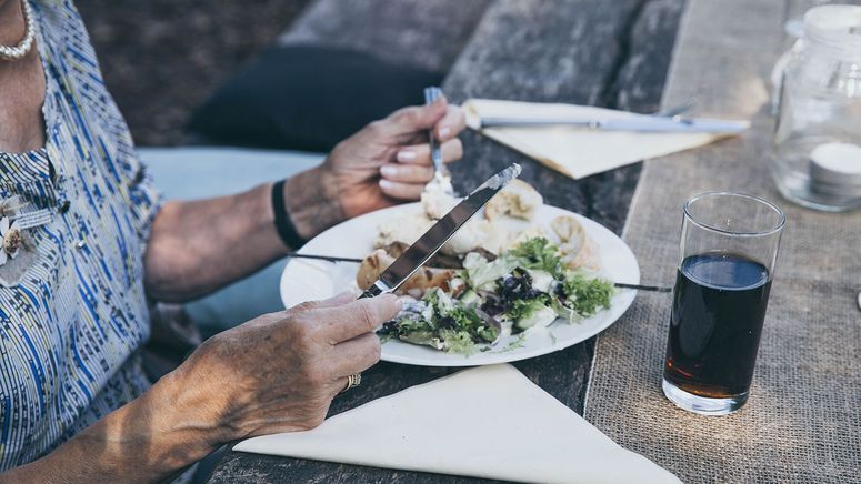Diversity in residential care- photo of am older woman's hands holding a knife and fork over a plate of salads, sitting at a table. 