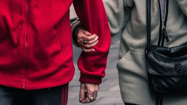 Residential aged care- cropped photo of an older couple's torsos walking, with arms linked, One is wearing a red jacket and the other grey with a black shoulder bag