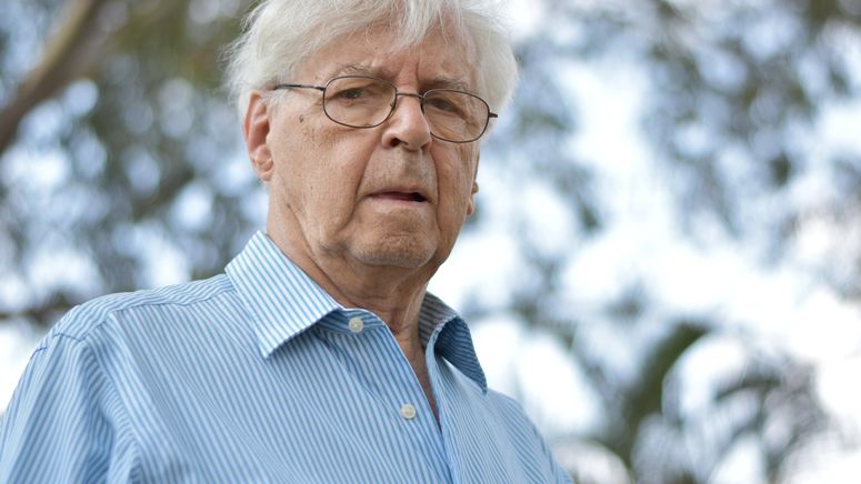 Photo of an older man with white hair and glasses looking down at the camera no smiling. He wears a blue business shirt.