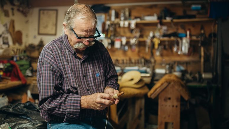 Photo of an older man balding with grey hair a moustache and glasses working on something with his hands in a workshop or shed