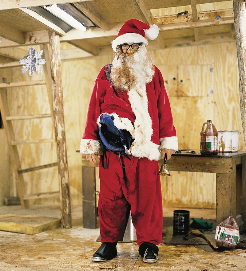 Paul McCarthy dressed as Santa standing on a plywood set in front of a table