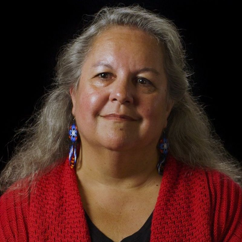 portrait of woman with long grey hair, wearing a red knitted cardigan and blue beaded earrings