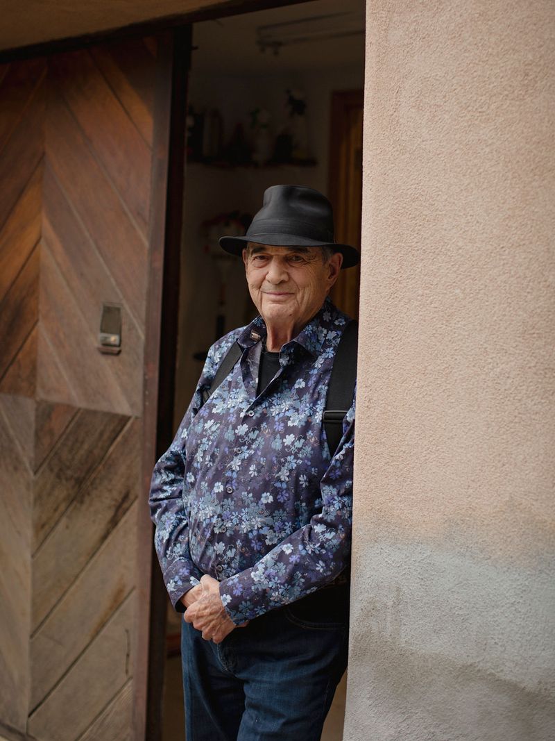 Artist Larry Bell wearing a hat and smiling while leaning against an entrance to his studio