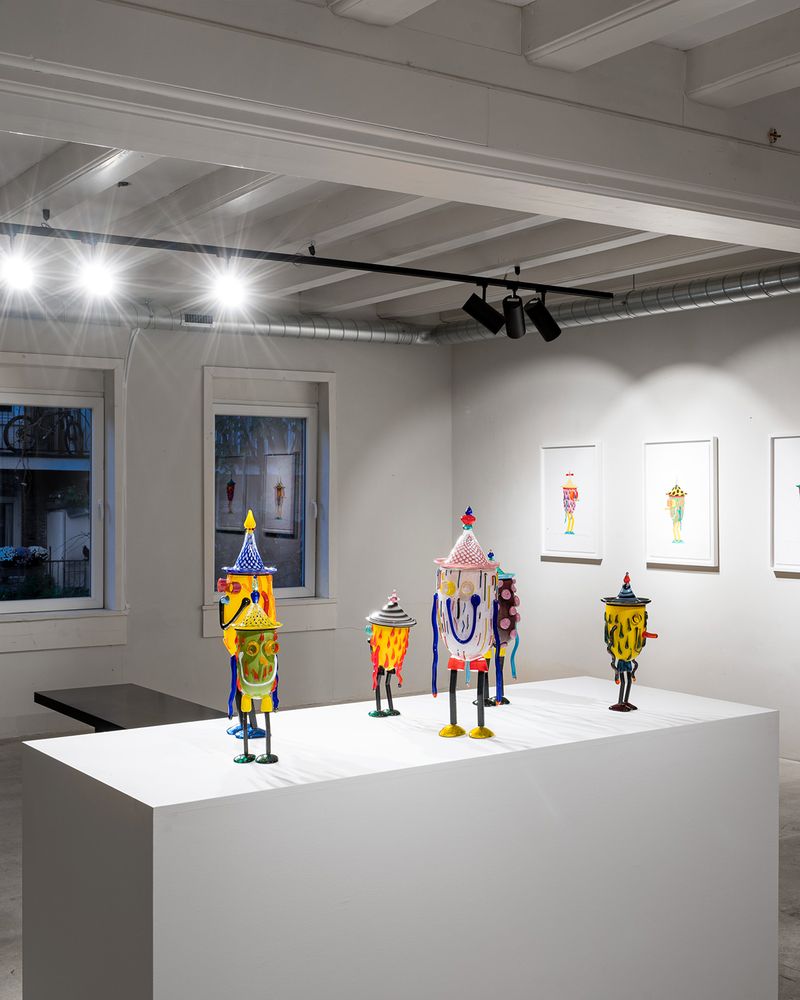 six smiling glass sculptures on white plinth with preparatory drawings framed on the wall behind