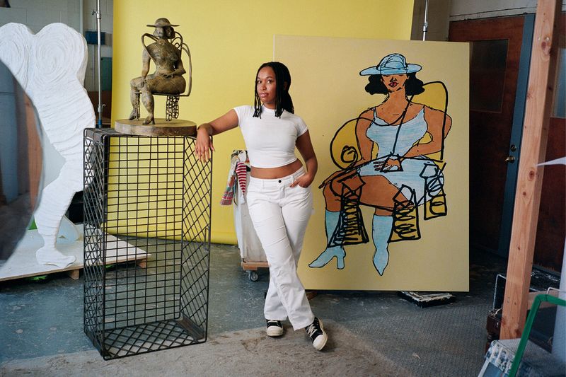 Tschabalala Self in her New Haven studio, wearing white and posing with a bronze sculpture and large yellow painting