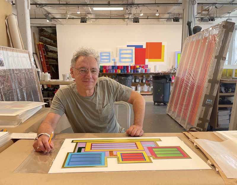 Peter Halley smiling at the camera while signing prints in his New York studio