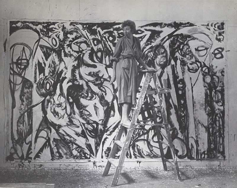 artist Lee Krasner wearing a robe and standing on a ladder next to a large painting 