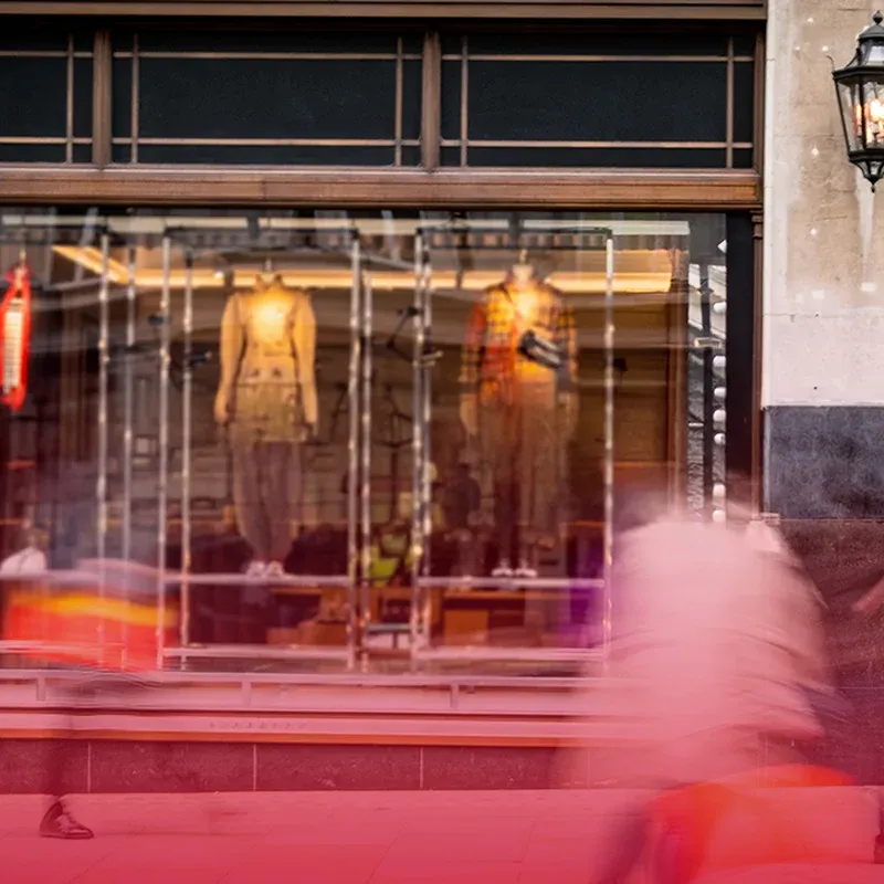 Square photo of a retail storefront at dusk with passersby blurred in motion outside the lit shop windows