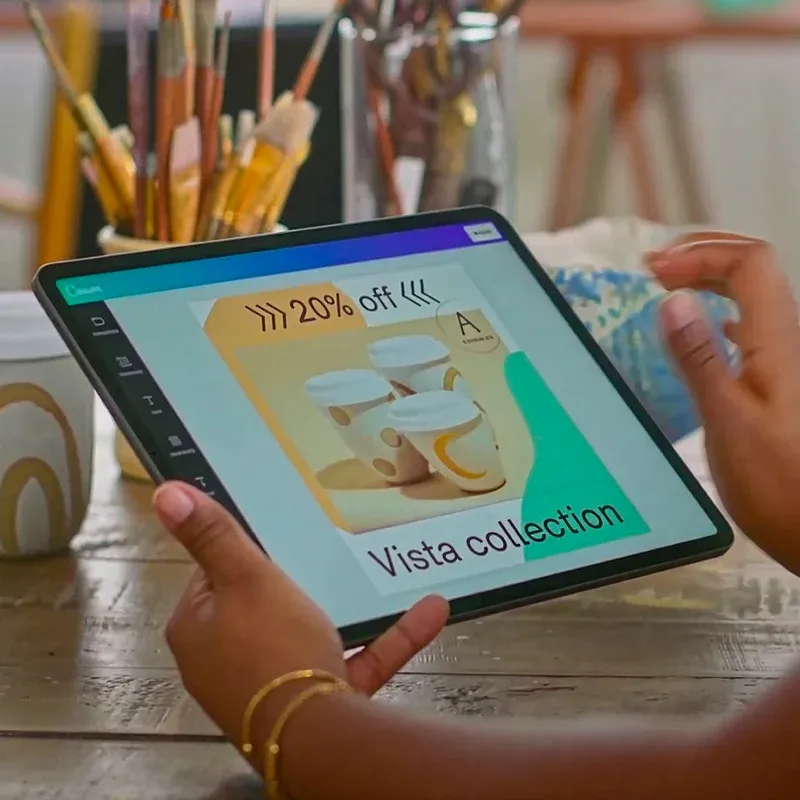 Hands using the Canva app on a tablet at a desk beside a coffee cup