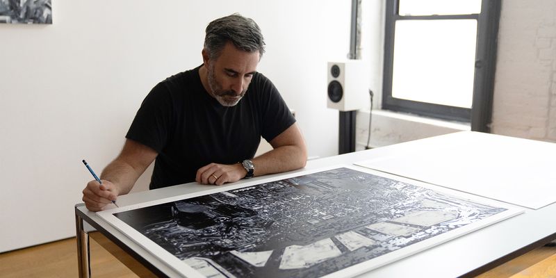 artist sitting behind table, signing a large monochromatic screenprint