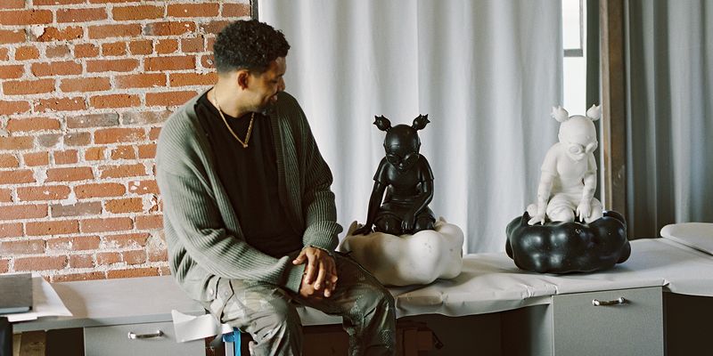 Artist in khaki green cardigan sitting on a table next to two of his marble sculptures