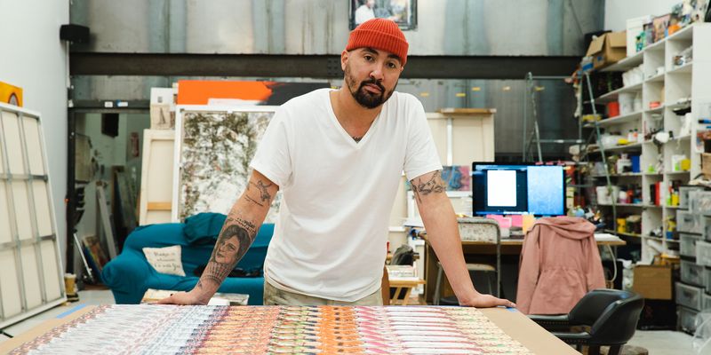 artist posing for a photo behind a table on which his prints are laid out – both of his hands are resting on the edge of the table