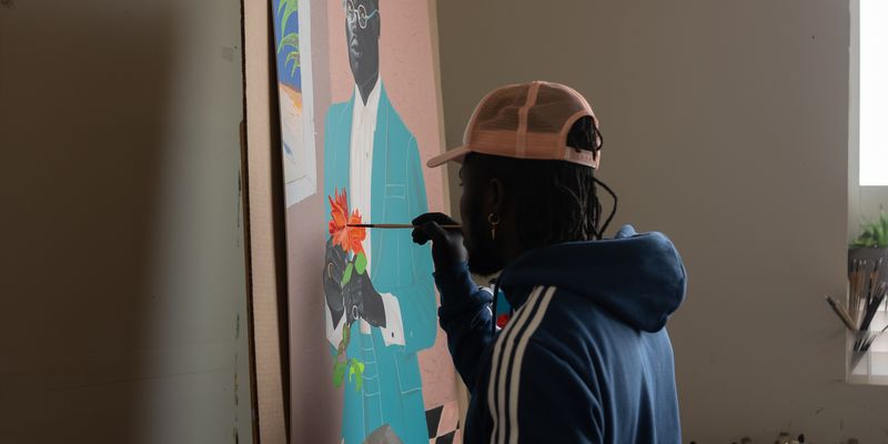 artist Otis Kwame Kye Quaicoe hand holding a paintbrush up to a painting to add detail to an orange flower