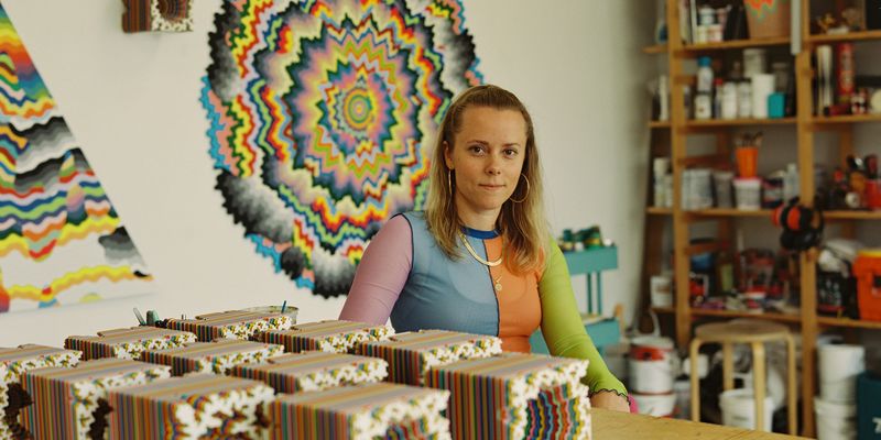 Jen Stark sat behind table of her sculptures in her studio with painting behind her on wall